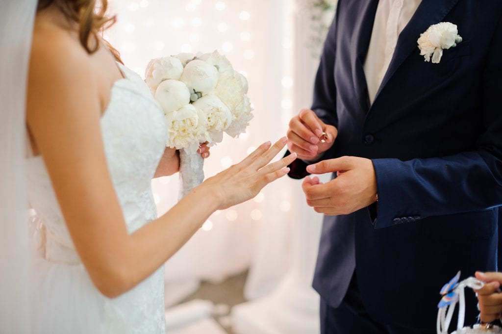 תרגום תעודת נישואין: בתמונה: חתן בחליפה כחולה עונד טבעת נישואין מזהב על אצבעה של כלה בשמלה לבנה